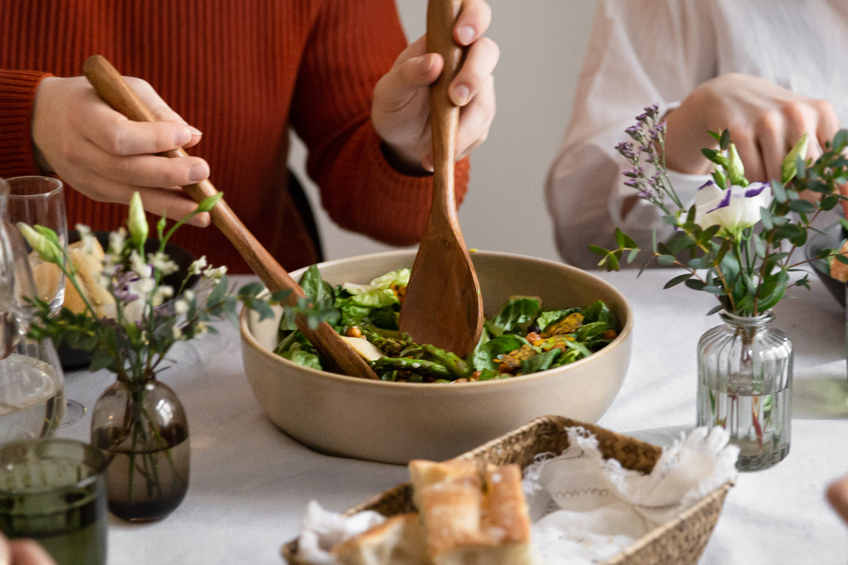 Lounaan kattaus, jossa henkilö ottaa salaattia kulhosta, pöydällä myös leipää, vesikannu ja koristekukkia.