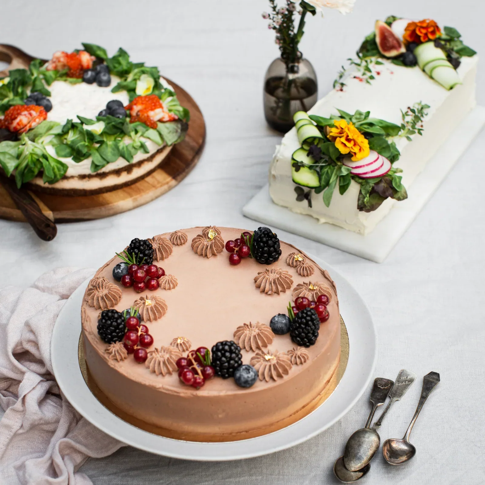 Juhlava kakkukattaus, pellavaliina ja hopealusikat, kesäkukilla ja marjoilla koristeltuja voileipäkakkuja ja suklaakakku.