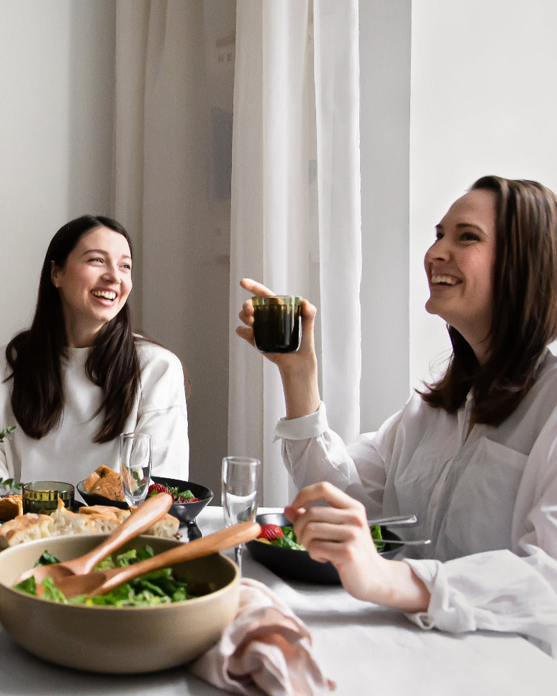 Rento lounashetki vaalein sävyin sisustetussa ruokasalissa, hymyilevät ystävykset, pöydässä katettuna salaattia, foccacciaa ja kuohuviiniä.