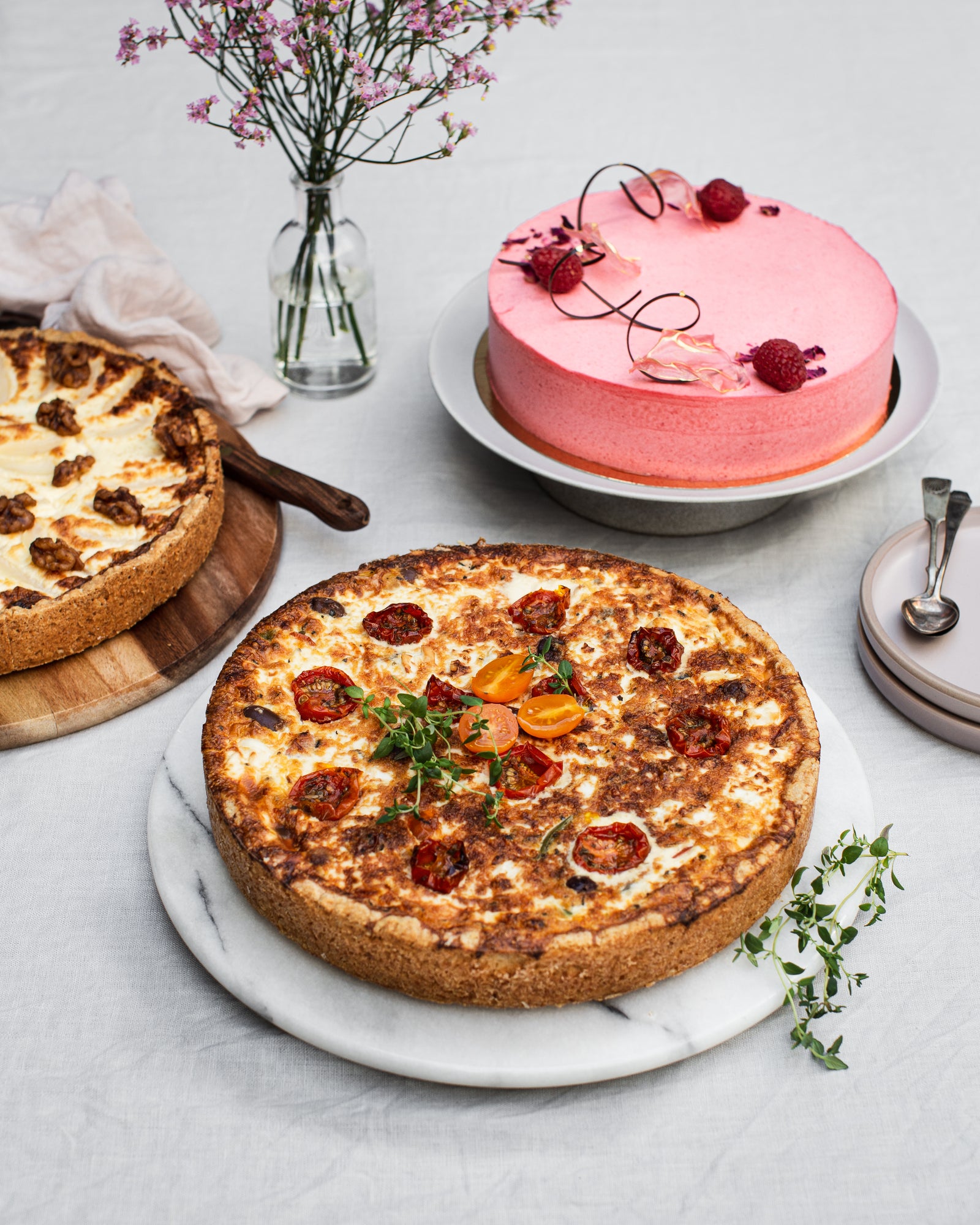 Juhlava kattaus pöydällä, jossa marjainen juustokakku ja kaksi suolaista piirakkaa aseteilla. 