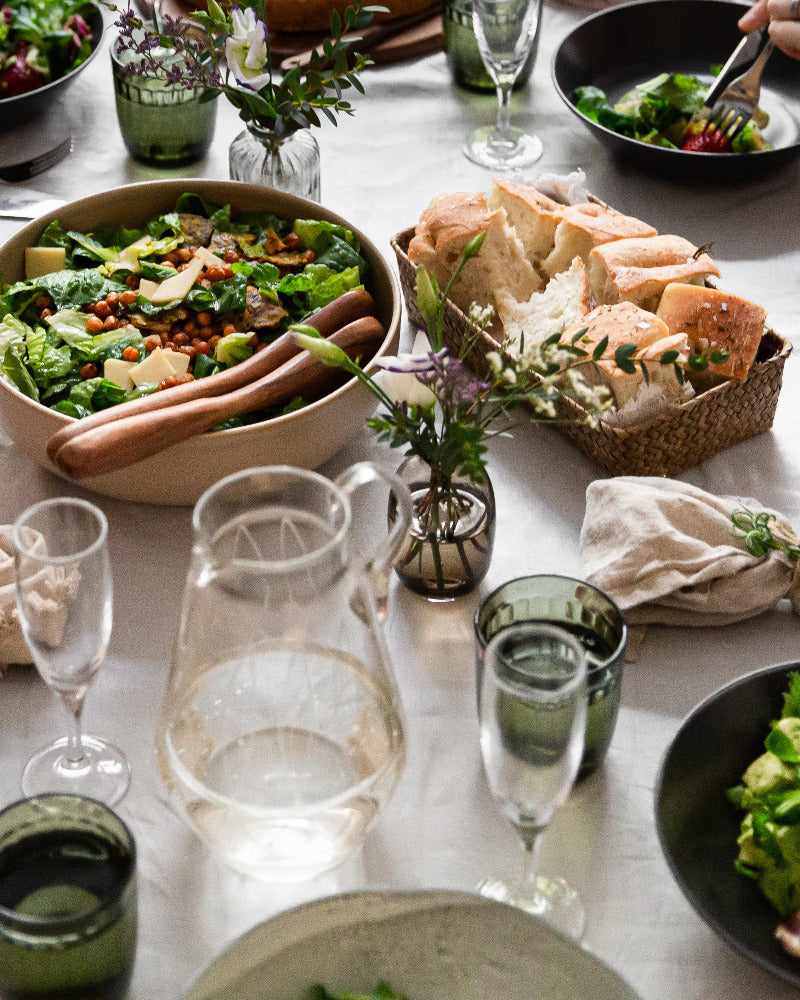 Runsas lounaspöytä katettuna valkoiselle pellavaliinalle, salaatteja ja foccacciaa, kuohuviinilasit ja niittykukkia.