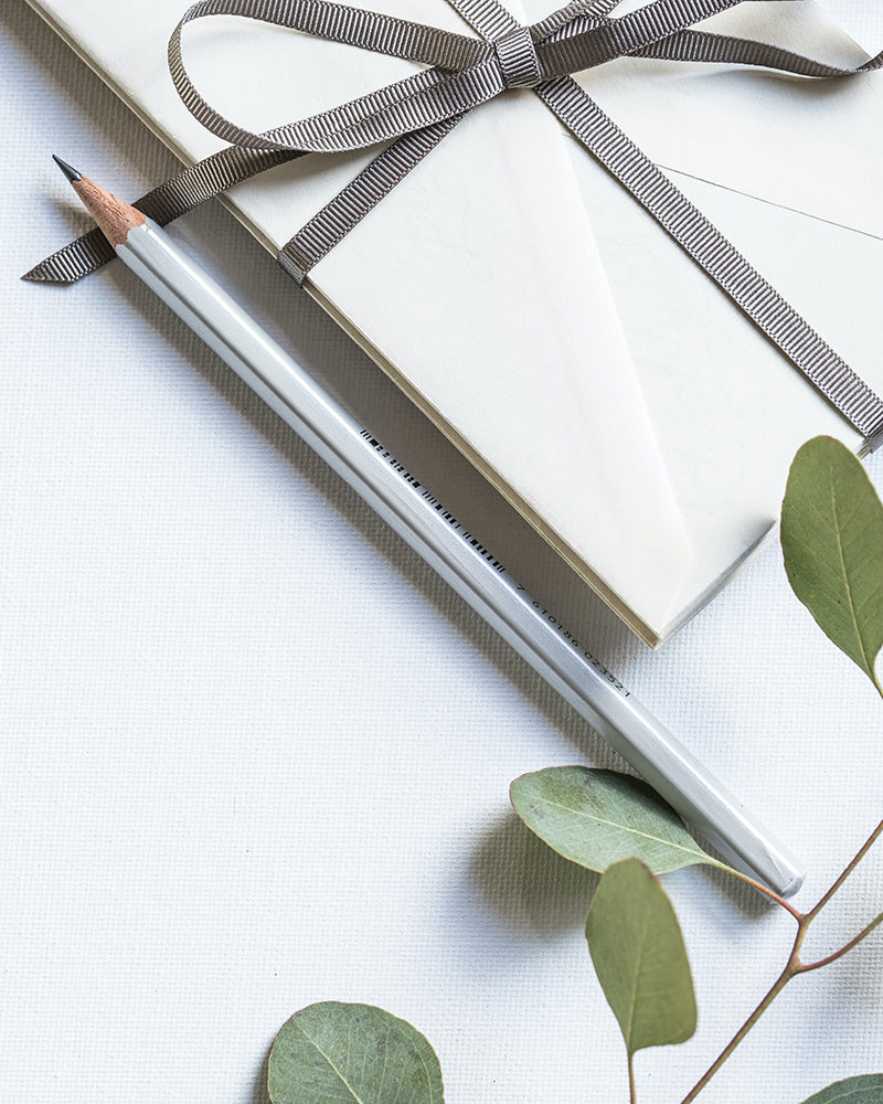 Rusetilla koristeltu kirjekuori ja lyijykynä koivunoksan vieressä pöydällä. 